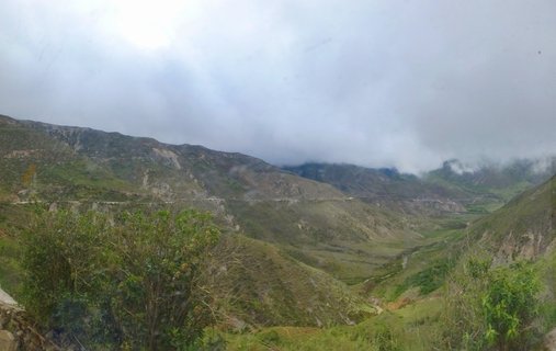 Green mountain valley with a winding road visible on the hillsides, clouds covering the mountain peaks under overcast sky