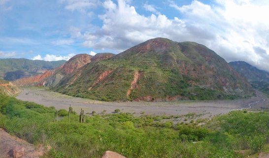 Mountain valley landscape with green vegetation, tall cacti, and red rock mountains under blue sky with white clouds