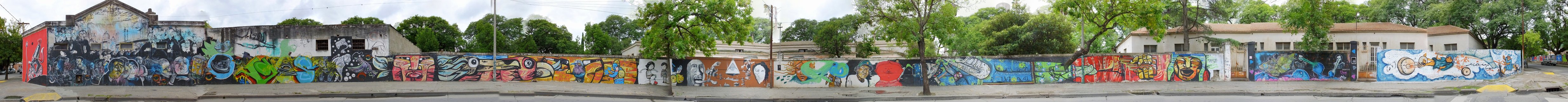 Long panoramic street view showing a concrete wall covered in colorful graffiti murals with buildings and trees in the background