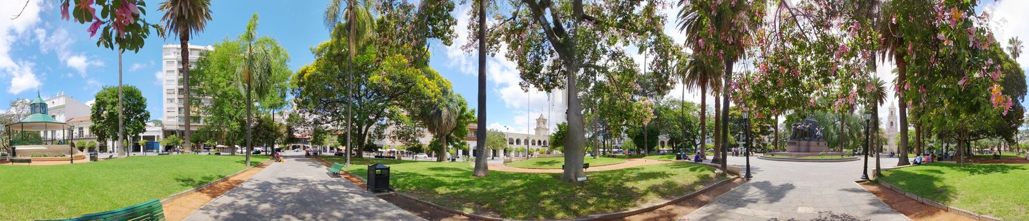 Wide panoramic view of a tree-filled urban plaza with cathedral towers, palm trees, green lawns, walkways, benches with people, and buildings under a blue sky with clouds in Salta, Argentina