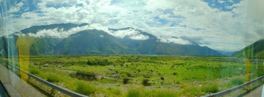 Panoramic view of green mountain valley in Chile with cloud-covered peaks, guardrail visible in foreground, taken from vehicle window