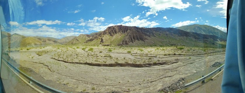 Desert landscape with brown stratified mountains under blue sky with white clouds, sparse green shrubs on arid ground, viewed from vehicle window with guardrail visible