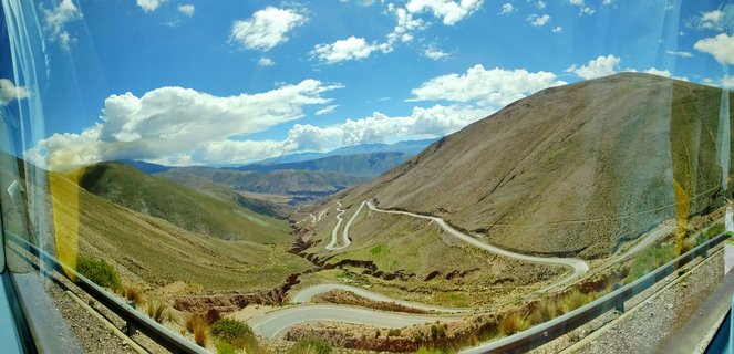 A winding paved road with multiple switchbacks curves through brown and green arid mountain hills under a blue sky with white clouds, viewed from inside a vehicle