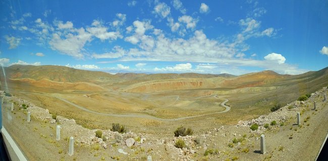 Panoramic landscape showing a winding road through brown and ochre colored desert hills under a blue sky with white clouds in the Atacama Desert, Chile