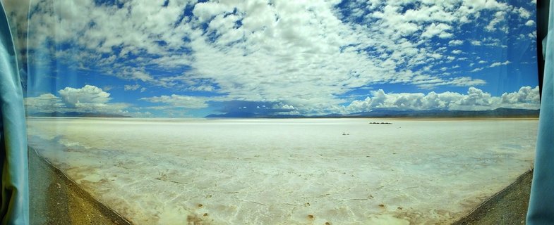 Panoramic landscape showing a vast white salt flat extending to distant mountains under a blue sky with white clouds, viewed through a vehicle window with blue curtains visible on both sides