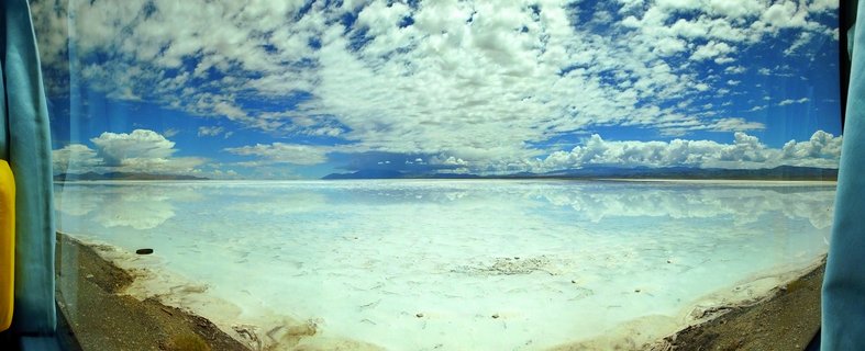 Vast salt flat landscape with mirror-like water reflections showing dramatic blue sky with white cumulus clouds, distant mountain ranges on horizon, white salt crust patterns in foreground, Salar de Atacama, Chile