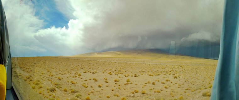Desert landscape showing sandy terrain with scattered small shrubs under a dramatic sky with large storm clouds approaching over distant hills