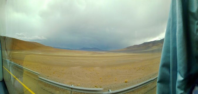 Desert landscape with brown arid plains, distant mountains, and dark storm clouds viewed through a vehicle window with visible guardrail and blue curtain