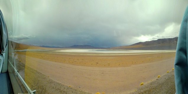 Desert landscape showing a white salt flat in the middle distance, brown arid terrain with sparse yellow grass tufts, dark storm clouds overhead, and mountains on the horizon, photographed from inside a vehicle with window reflections visible at the edges