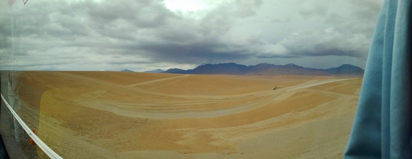 Desert landscape with golden-brown terrain, dirt roads, distant mountains, and cloudy overcast sky viewed from inside a vehicle