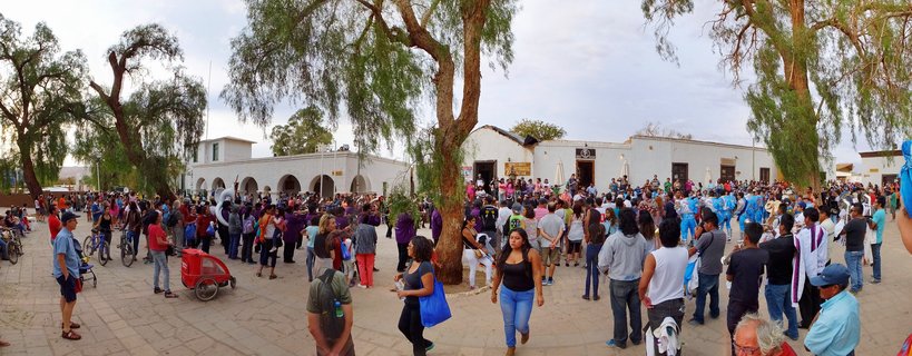 Large crowd of people gathered in a town plaza with white colonial buildings, large trees, and costumed performers during a festival in San Pedro de Atacama, Chile