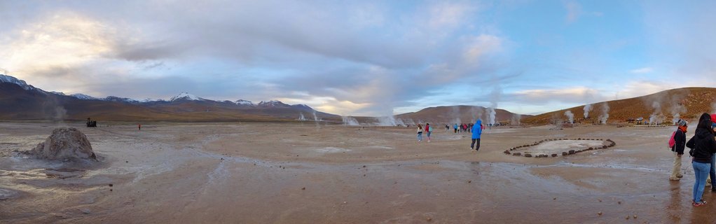 Panoramic view of El Tatio Geothermal Field in Chile's Atacama Desert at dawn, showing multiple white steam plumes rising from geothermal vents across barren brown terrain, with tourists in warm clothing walking among the geysers and snow-capped Andes mountains visible in the distant background under a partly cloudy sky