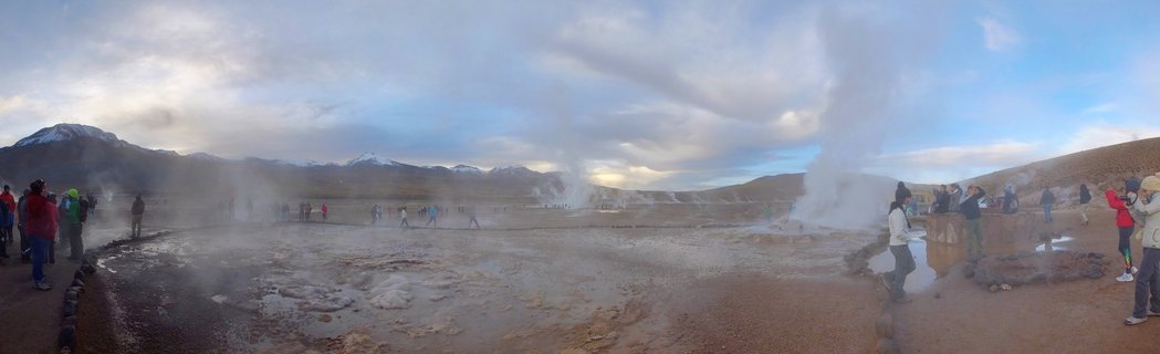 Panoramic view of El Tatio geysers with tourists walking among steam vents, snow-capped mountains in background, Atacama Desert, Chile