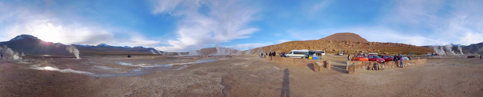 Panoramic view of El Tatio geyser field showing steam vents across desert terrain with mountains, tour buses, and visitors in early morning light