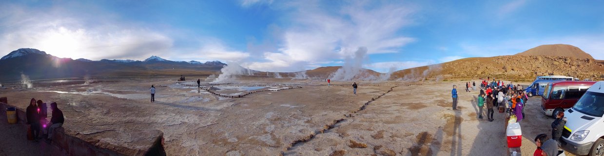 Panoramic view of El Tatio geysers in Chile's Atacama Desert showing multiple steam plumes rising from geothermal vents with tourists and tour buses visible, snow-capped mountains in background under blue sky at sunrise
