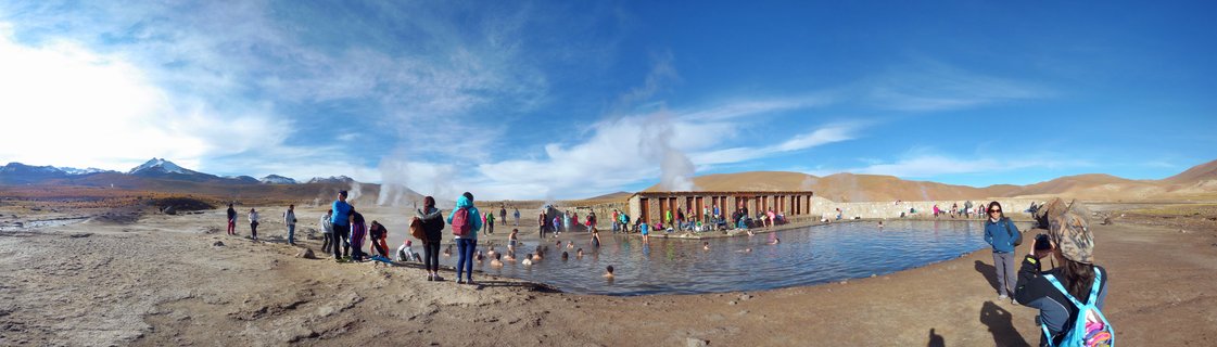 People bathing in outdoor geothermal hot spring pools with steam rising, wooden facilities, desert mountains and blue sky in San Pedro de Atacama, Chile