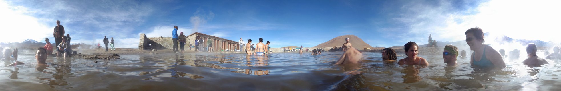 People bathing in natural thermal hot springs with steam rising, desert mountains and blue sky in background at Termas de Puritama, Chile