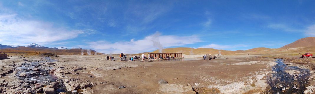 Panoramic view of El Tatio geothermal field showing steam rising from vents, visitors on wooden platforms, arid brown terrain with streams, and snow-capped mountains under blue sky