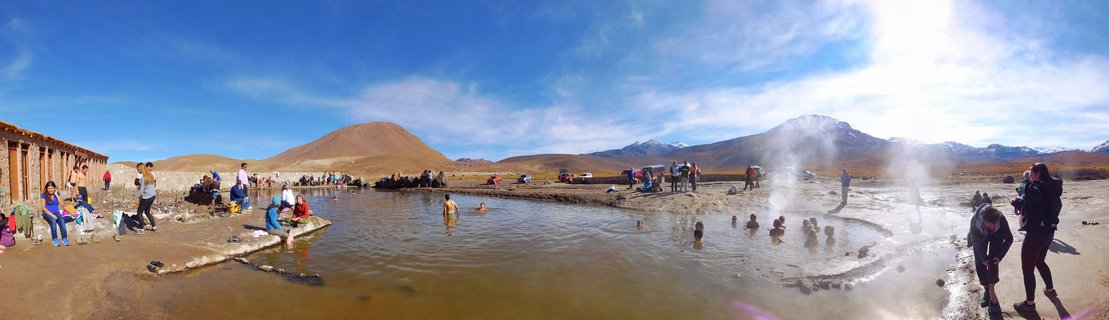 People bathing in outdoor thermal hot springs with steam rising, desert mountains and blue sky in background at San Pedro de Atacama, Chile