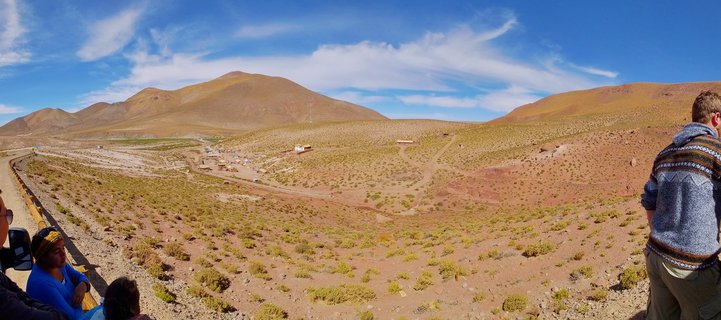 Wide desert landscape showing reddish-brown terrain with sparse green shrubs, barren mountains in the background, blue sky with white clouds, and people visible at the edges of the frame at a viewpoint