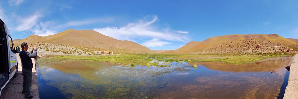 Panoramic landscape of San Pedro de Atacama wetlands showing a photographer with a white van on the left, calm reflective water in the foreground, green marshland vegetation, and arid desert mountains under a blue sky with white clouds in Chile