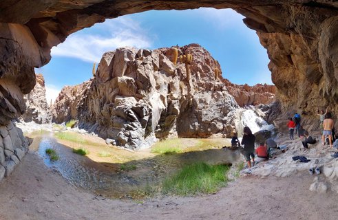 View from inside a cave looking out at a thermal waterfall and hot spring pool in the Atacama Desert, with tourists sitting by the water and tall cacti growing on rocky formations under a blue sky