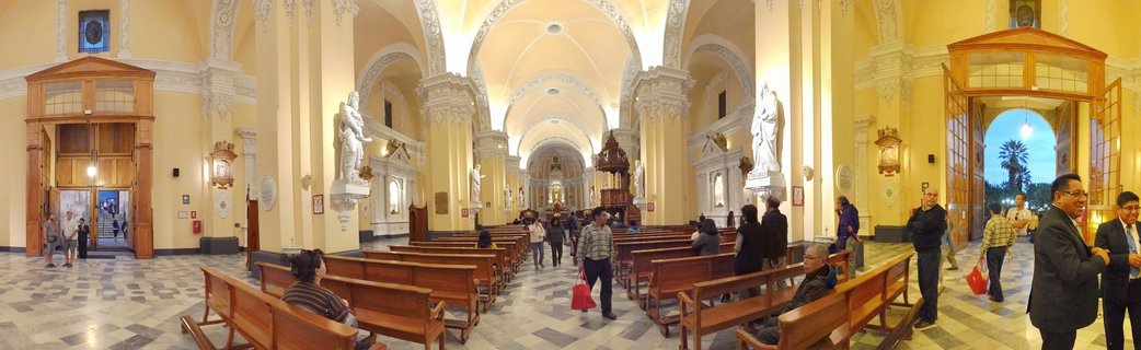 Interior view of a large cathedral with cream-colored walls, white ornate columns, wooden pews, and visitors walking through the nave