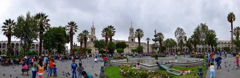 Wide view of a bustling colonial plaza with a white stone cathedral featuring two bell towers, surrounded by arcaded buildings, palm trees, a central fountain, and numerous people walking among pigeons