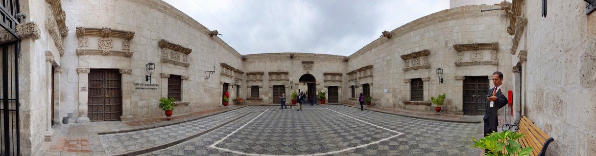 Wide courtyard surrounded by white stone colonial buildings with ornate windows, wooden doors, and people walking on patterned cobblestone ground under overcast sky