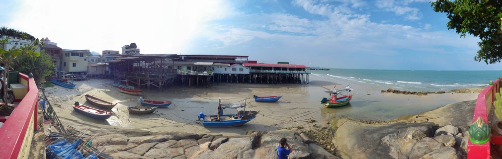 Panoramic coastal scene showing multiple colorful fishing boats on exposed sand at low tide with wooden stilt pier structures, buildings, and ocean under blue sky with scattered clouds in Hua Hin, Thailand