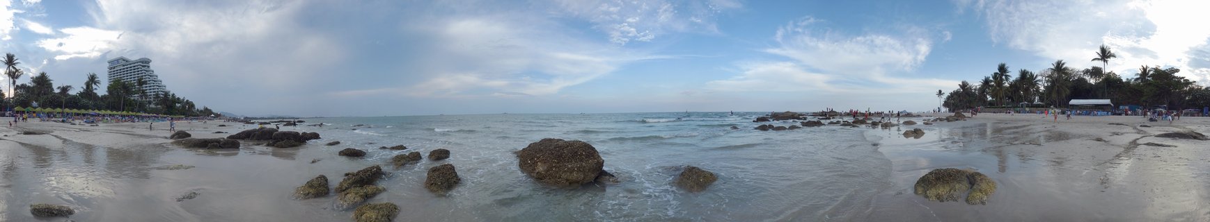 Wide panoramic beach scene showing rocky shoreline with large boulders, calm ocean water, sandy beach with palm trees, resort building in distance, blue sky with clouds