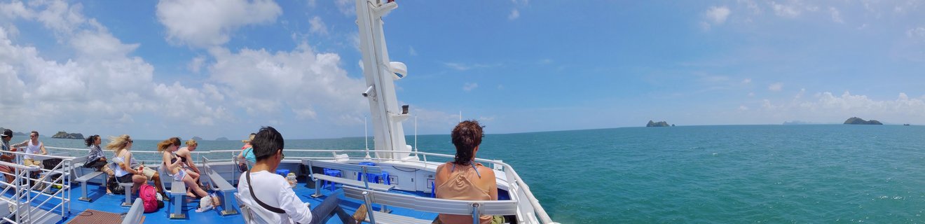 Passengers sitting on ferry boat deck with blue ocean water, white boat structure, distant islands, and cloudy blue sky in Thailand
