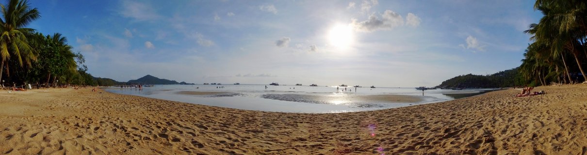 Wide panoramic beach scene showing golden sand with footprints, calm shallow water, multiple boats in the distance, palm trees on both sides, people on the beach, hills in the background, and bright sun in blue sky with scattered clouds