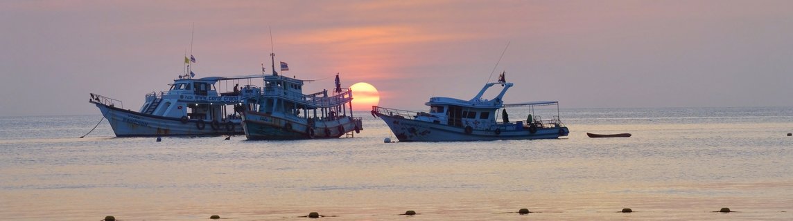 Three blue Thai fishing boats anchored in calm ocean water with orange sunset sun visible between the vessels