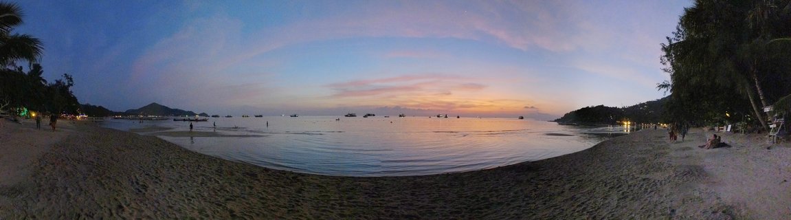 Wide panoramic beach scene at dusk showing calm bay water with boats, palm trees on both sides, people on the sand, colorful sunset sky with orange and blue colors, and hills in the background