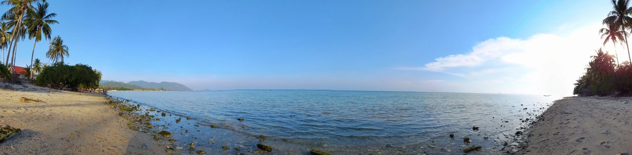 Wide panoramic view of a tropical beach in Thailand showing palm trees on both sides, calm blue ocean water, sandy shore with rocks, and distant mountains under a clear sky