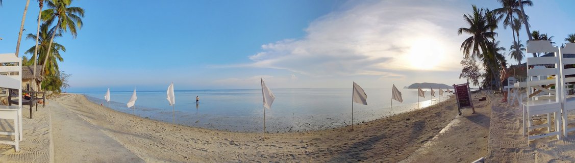 Panoramic beach scene at sunset showing white sand, calm ocean water, white flags on poles, palm trees on both sides, white wooden furniture, and a person standing in shallow water with distant island on horizon