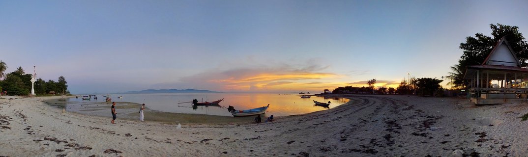 Panoramic beach scene at sunset showing multiple traditional Thai long-tail boats in calm water, sandy shore with people, tropical vegetation, and a pavilion structure with orange and yellow sky