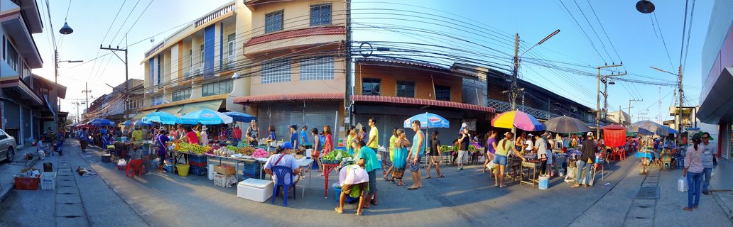 A wide street market scene in Thailand with vendors selling fresh fruits and vegetables under blue and multicolored umbrellas, shoppers browsing stalls, and two to three-story buildings lining the paved street under a clear blue sky