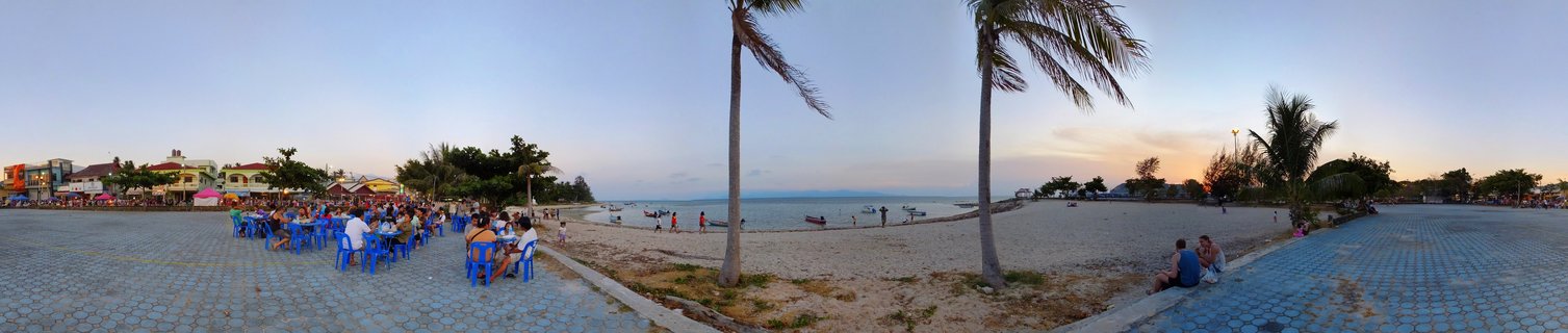Panoramic view of beachfront restaurants with tourists dining at blue tables, palm trees, sandy beach, and sunset over the ocean in Koh Phangan, Thailand