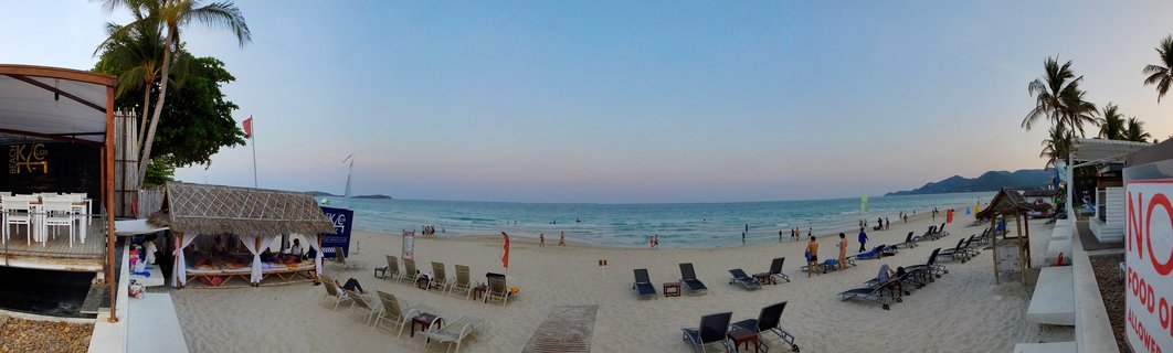 Panoramic view of a tropical beach at dusk showing white sand, turquoise ocean, palm trees, beach lounge chairs, thatched cabanas, beachfront restaurants, and distant mountains under a clear gradient sky
