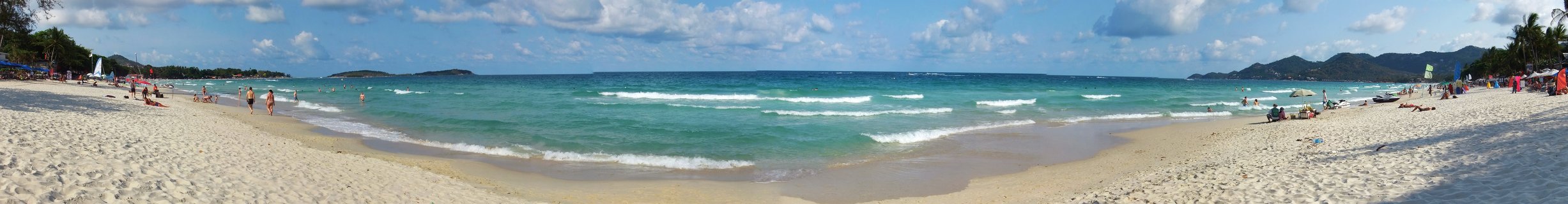 Wide panoramic photograph of a tropical beach in Thailand showing white sand, turquoise ocean with waves, people walking and relaxing on the shore, distant islands on the horizon, blue sky with clouds, and palm trees along the coastline