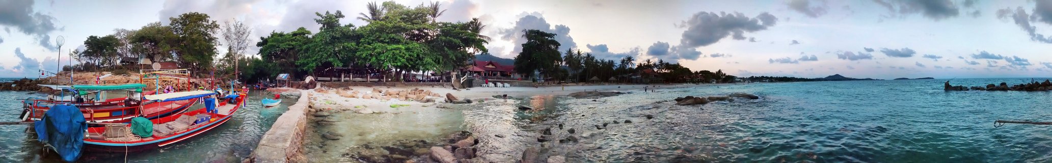 Panoramic view of colorful Thai longtail boats docked at a pier with tropical beach, palm trees, resort buildings, and turquoise ocean water under cloudy evening sky in Koh Samui, Thailand