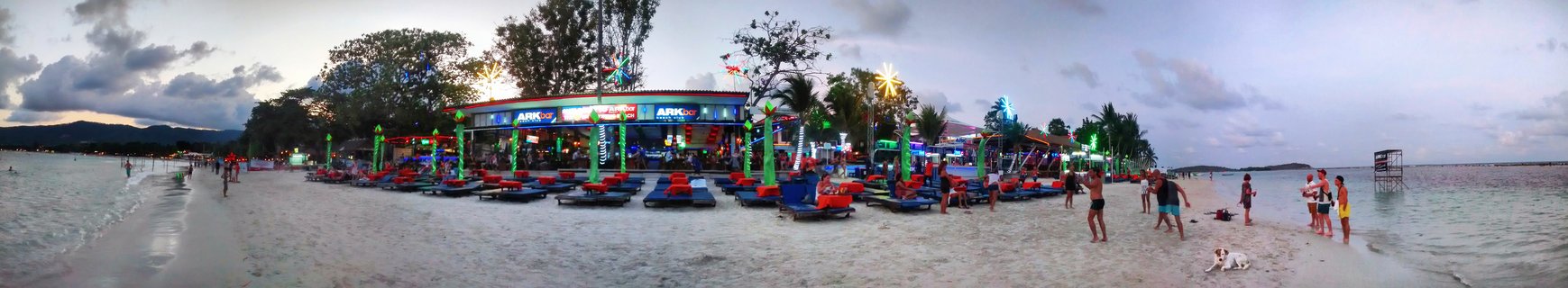 Panoramic view of a beach bar with bright neon lights, tourists on white sand beach, lounge chairs, palm trees, and ocean at twilight in Koh Samui Thailand
