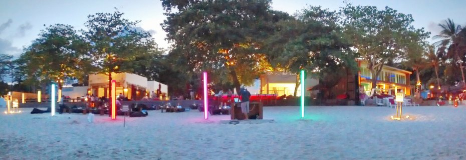 Wide beach scene at dusk showing a beach bar with colorful vertical LED light tubes in cyan, red, pink and green, with illuminated buildings, tropical trees and people on beach loungers on white sand