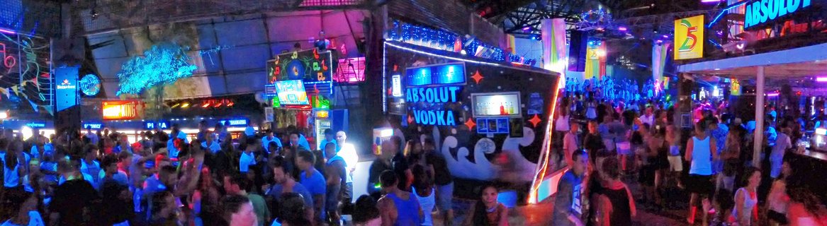 Large crowd of people at a nightclub with neon blue and purple lighting, bar areas, and Absolut Vodka signage visible in Koh Samui, Thailand