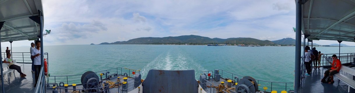 View from the back deck of a ferry showing passengers on both sides, the vessel's wake in turquoise water, and a mountainous tropical island coastline in the distance under a cloudy sky