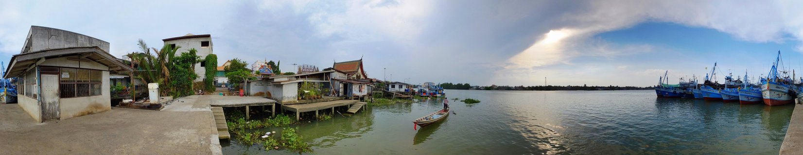 Panoramic waterfront scene showing traditional wooden stilt houses on the left, a person in a small boat in the center, and multiple blue fishing boats docked on the right side of a harbor in Thailand