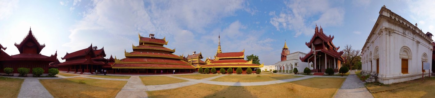Panoramic view of Mandalay Royal Palace showing traditional Burmese red wooden buildings with golden tiered roofs and spires, white colonial structures, paved walkways, and manicured lawns under blue sky with clouds