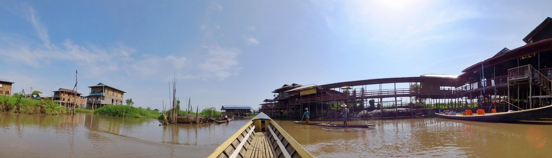 View from a wooden boat on Inle Lake showing traditional stilt houses, local fishermen in small boats, and wooden walkways under a blue sky
